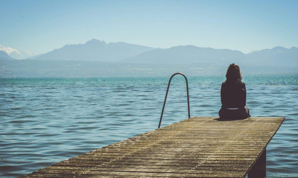 a woman sits on the end of a dock during daytime staring across a lake